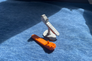 An orange, plastic lego tool sits next to a replica cast in pewter with its sprue still attached