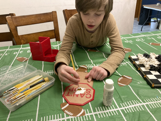 A person painting the stencil on the back panel of the Can't Stop dice tower.  The words "Can't Stop" are painted in white onto red acrylic.